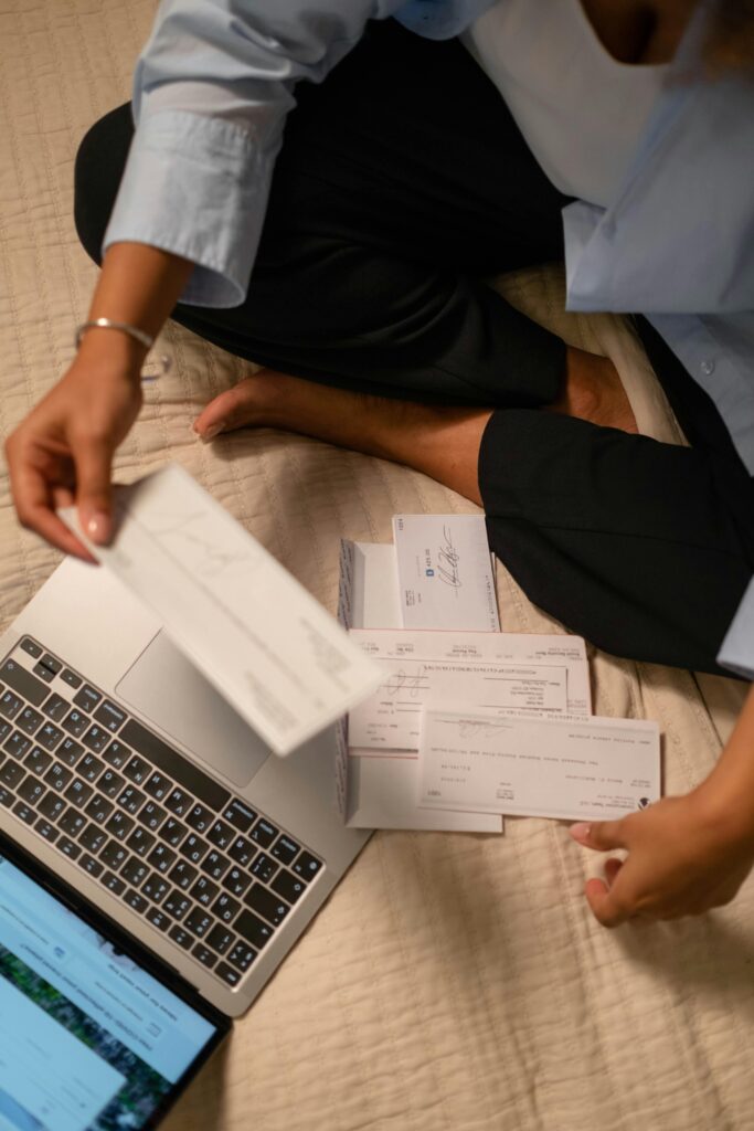 An adult organizing cheques and using a laptop indoors for financial tasks.