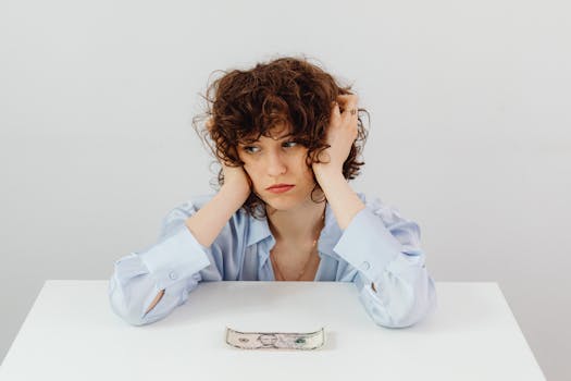 A woman in a blue blouse looks worried with a dollar bill on a table, symbolizing financial stress.