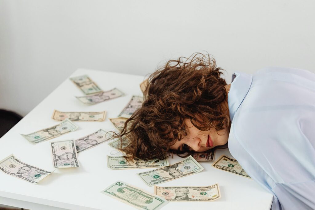 A woman with curly hair resting her head on a table covered in US dollar bills.