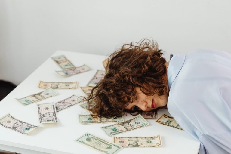 A woman with curly hair resting her head on a table covered in US dollar bills.